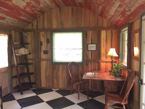 Cozy wooden interior with a table, two chairs, a lamp, and a shelf, featuring a checkered floor and natural light.