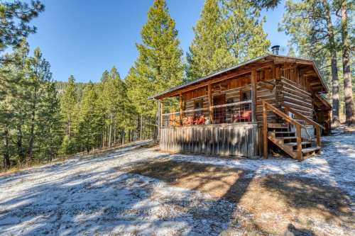 A rustic log cabin surrounded by tall pine trees and a snowy path, set against a clear blue sky.