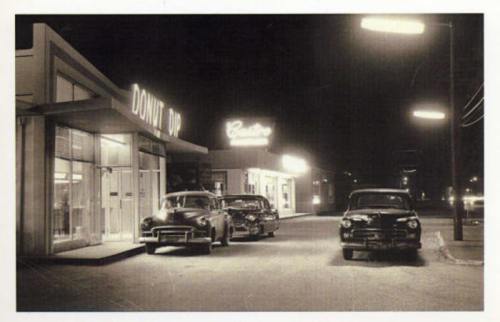 A vintage night scene featuring classic cars parked outside a diner and a brightly lit restaurant.