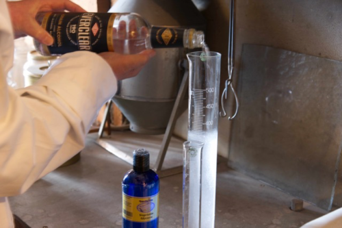 A person in a lab coat pours a clear liquid from a bottle into a graduated cylinder on a workbench.