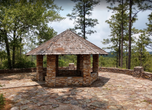 A stone gazebo with a wooden roof, surrounded by trees and a scenic view of rolling hills.