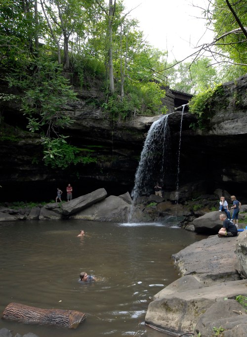 A serene waterfall cascades into a pool surrounded by rocks and lush greenery, with people enjoying the scene.
