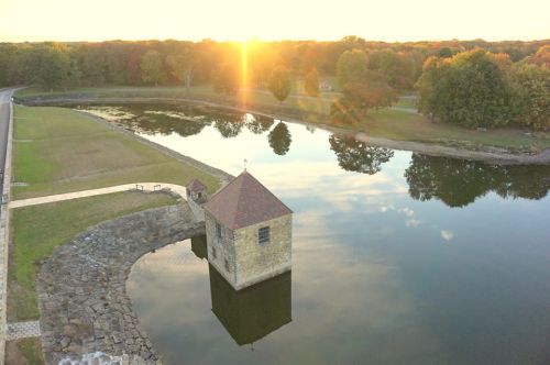A serene sunset over a calm lake, reflecting a small stone building and surrounding trees in autumn colors.