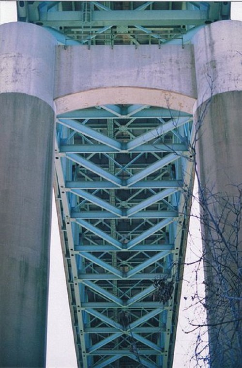 View from underneath a bridge, showcasing its blue steel structure and concrete pillars against a clear sky.