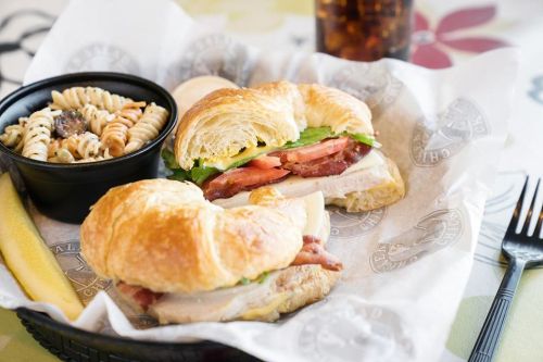 A croissant sandwich filled with turkey, lettuce, and tomato, served with a side of pasta and a drink.