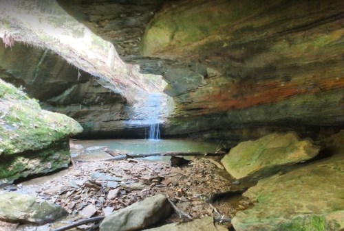 A serene cave scene with a small waterfall cascading into a pool, surrounded by mossy rocks and earthy tones.