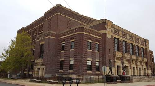 Historic brick building with decorative elements, featuring large windows and a statue in front, set against a cloudy sky.