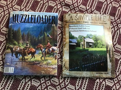 Two magazines on a patterned surface: "Muzzleloader" featuring horseback riders and "A Simple Life" showcasing a cabin.