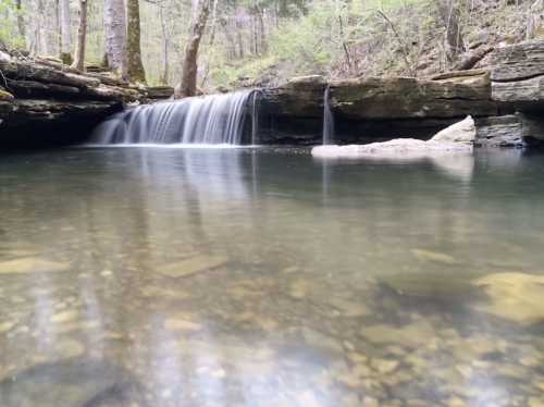 A serene waterfall cascades into a calm pool, surrounded by lush greenery and rocky terrain.