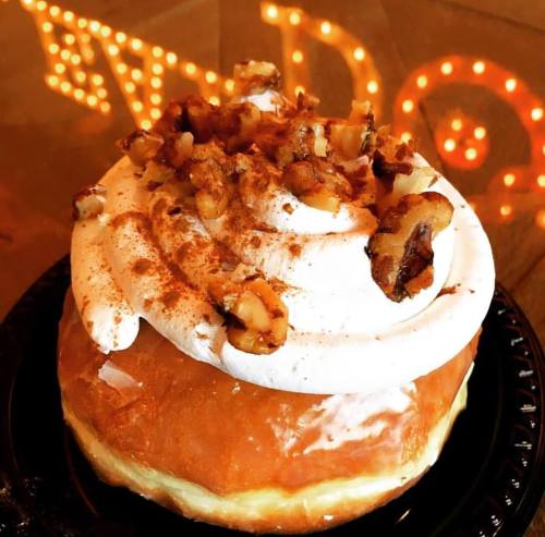 A frosted donut topped with whipped cream, walnuts, and a sprinkle of cinnamon on a black plate.