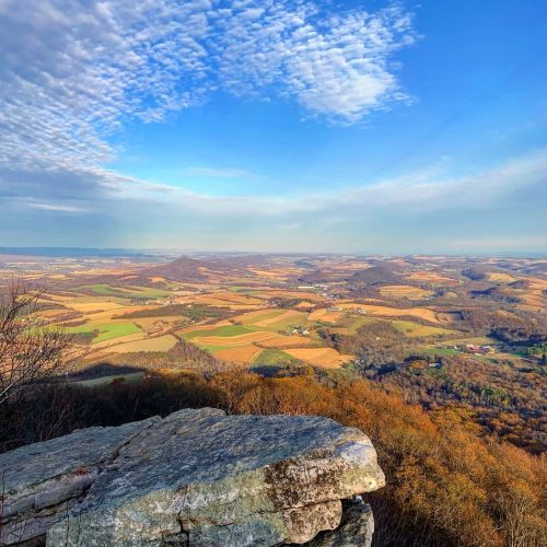 A panoramic view of rolling hills and fields under a blue sky with scattered clouds, seen from a rocky overlook.