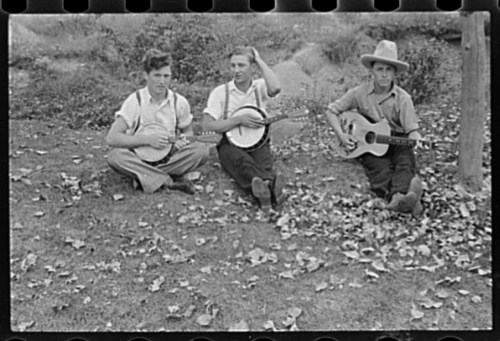 Three young men sitting on the ground with instruments, surrounded by fallen leaves, enjoying a musical moment outdoors.