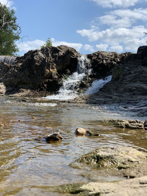 A small waterfall cascades over rocky terrain into a calm stream under a blue sky with scattered clouds.