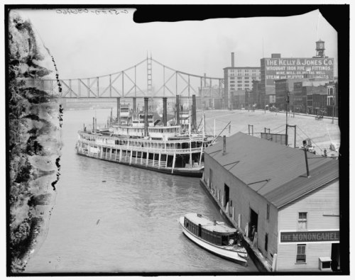 Historic river scene featuring a steamboat docked by a warehouse, with a bridge and buildings in the background.