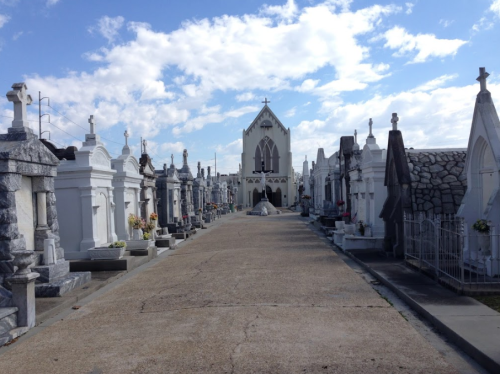 A cemetery pathway lined with ornate tombs and a church in the background under a blue sky with scattered clouds.