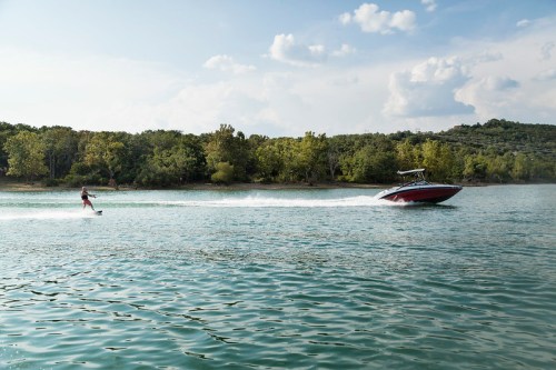 A person water skiing behind a boat on a calm lake, surrounded by green trees and a clear blue sky.