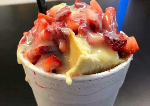 A cup filled with cake topped with strawberries and a creamy sauce, served with two straws.
