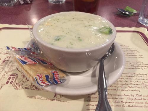 A bowl of creamy broccoli soup served with a spoon and a packet of crackers on a plate.