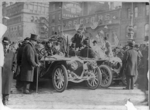 A historic black-and-white photo of a parade with people in top hats and vintage cars, decorated with flags.