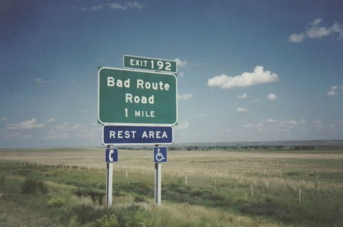 Green highway sign indicating "Bad Route Road" exit 192, with a rest area symbol, set against a blue sky and open fields.