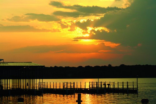 Sunset over a calm lake, with clouds and a silhouetted dock reflecting on the water's surface.