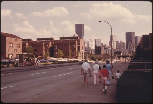 A group of people walking along a city street with buildings and cars in the background on a sunny day.