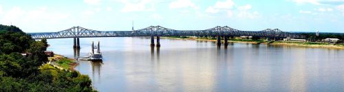 A steel bridge spans a wide river, with a boat docked nearby and lush greenery along the shore under a blue sky.