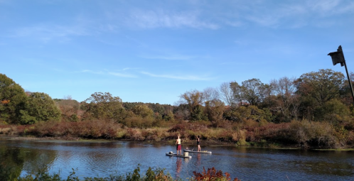 Two people stand on a small raft in a calm river, surrounded by autumn foliage and a clear blue sky.