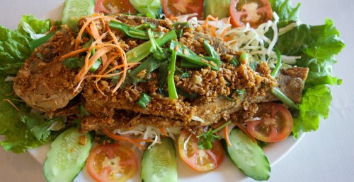 A whole fried fish garnished with herbs, vegetables, and crispy toppings, served on a bed of lettuce and sliced tomatoes.