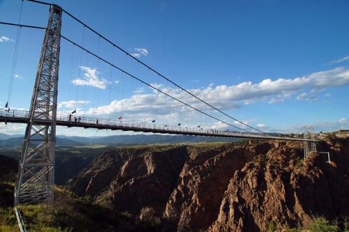 A suspension bridge spans a deep canyon, surrounded by mountains and a blue sky with scattered clouds.