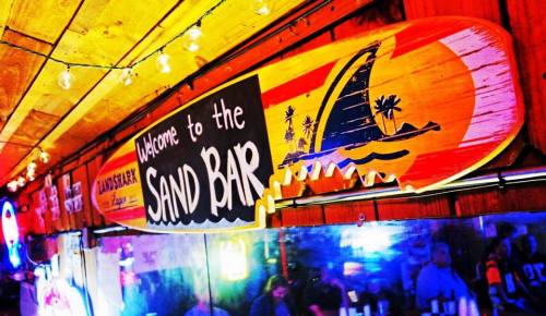 Colorful sign reading "Welcome to the Sand Bar" with a beach and sailboat design, hanging in a lively bar setting.