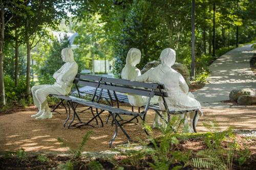 Four white sculptures of people sitting on a bench in a lush, green park setting.