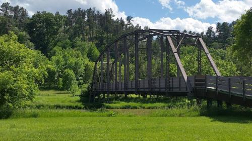 A metal arch bridge spans a lush green landscape with trees and a blue sky dotted with clouds.