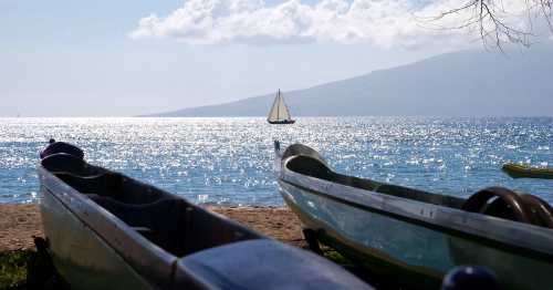 Two canoes on a sandy beach with a sailboat in the distance, sparkling water, and a scenic backdrop of hills.