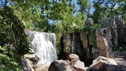 A serene waterfall cascades over rocks, surrounded by lush greenery and trees under a clear blue sky.