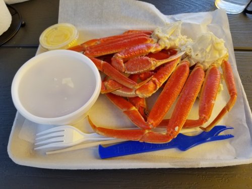 A tray with cooked crab legs, a container of dipping sauce, and plastic utensils.