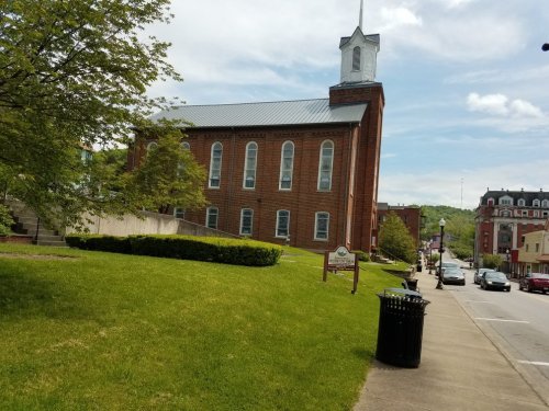 A brick church with a steeple, surrounded by greenery, on a sunny day with buildings and cars in the background.