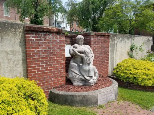 A stone sculpture of a seated figure holding a deer, surrounded by greenery and brick walls.