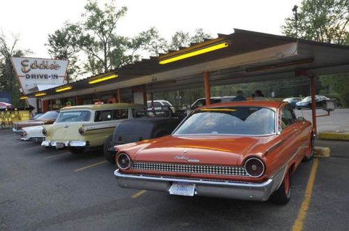 A row of vintage cars parked at Eddie's Drive-In, with a retro sign and trees in the background.