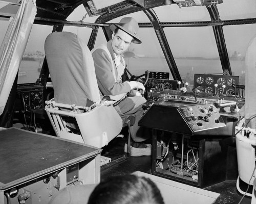 A man in a hat sits at the controls of a vintage aircraft cockpit, focused on flying.