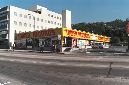 A colorful Tower Records store on a sunny day, featuring large posters and a parking lot in front.
