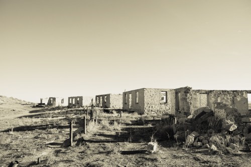 Deserted ruins of stone buildings with empty windows, set against a clear sky and barren landscape.