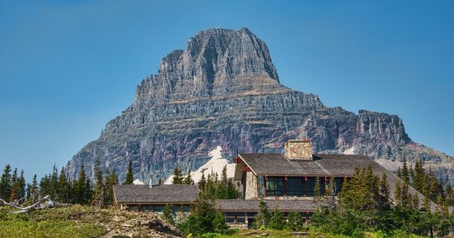 A large mountain towers over a rustic lodge surrounded by trees and greenery under a clear blue sky.