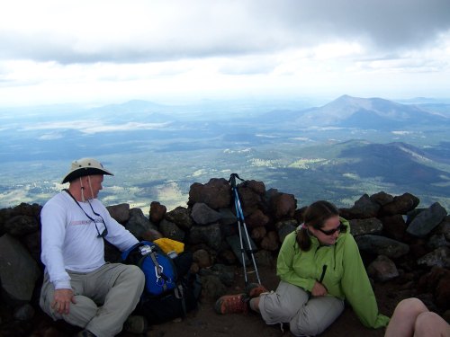Two hikers rest on a rocky mountain summit, overlooking a vast landscape of hills and valleys under a cloudy sky.