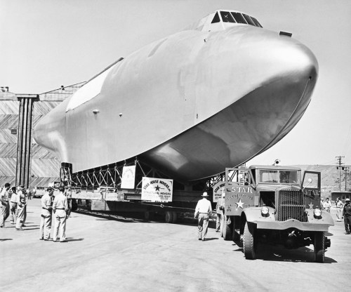 A large, futuristic aircraft on a trailer outside a hangar, surrounded by people and vintage trucks.