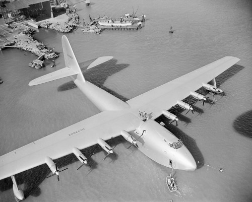 Aerial view of a large seaplane on water, with a dock and boats in the background. Black and white image.