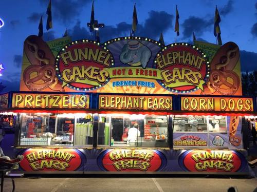 Colorful food stand at a fair featuring funnel cakes, elephant ears, pretzels, and corn dogs, with a vibrant night sky.