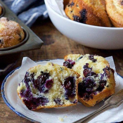 A plate with a halved blueberry muffin, revealing juicy blueberries, with more muffins in the background.