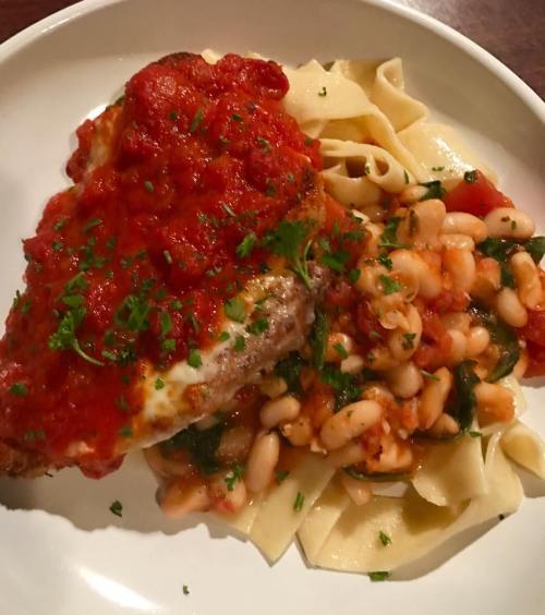 A plate of pasta topped with a breaded chicken breast and marinara sauce, served with white beans and herbs.