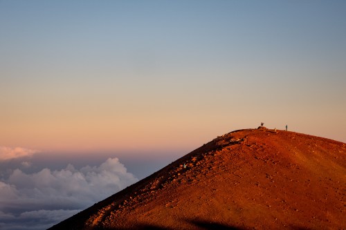 A barren mountain peak at sunset, with two small figures silhouetted against a colorful sky.